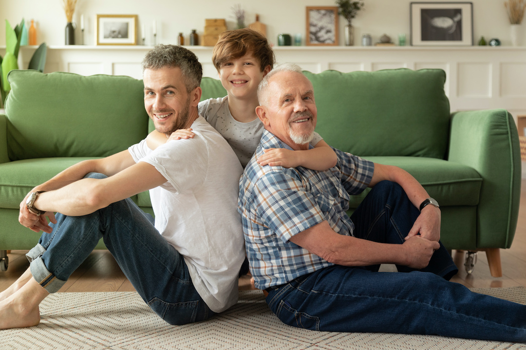 Happy multi-generational family father, son, grandfather hug each other sitting on floor in cozy home. Male generations